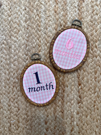 Hanging oval milestone discs reading '1 month' and '6 months' on gingham