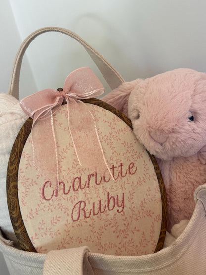 Pink floral oval baby name reveal sign with pink bow in a basket with plush lamb