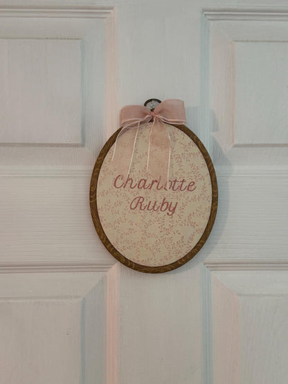 Pink floral embroidered baby name sign with pink bow in an oval hoop on a door