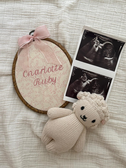 Pink floral oval baby name reveal sign with pink bow and plush bunny beside ultrasound cards